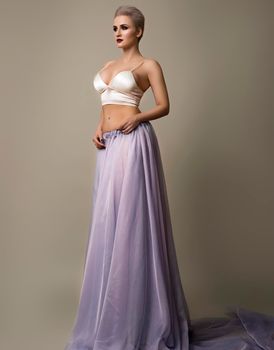 Young stylish woman with short haircut in white bra and transparent skirt posing on gray background.