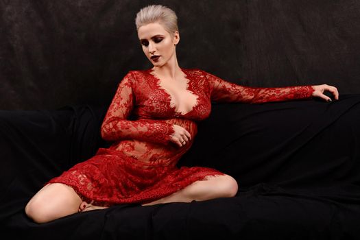 attractive short-haired young blond woman in red lace dress sits on the couch.