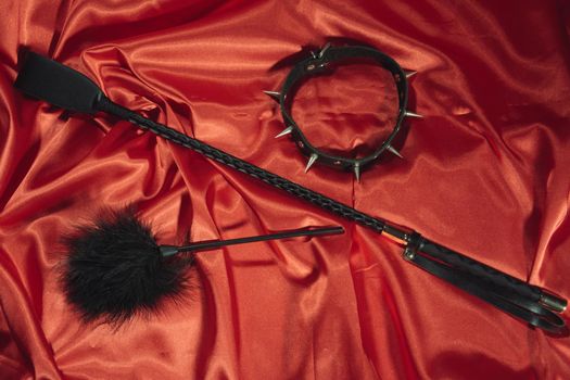 Bondage, kinky adult sex games, kink and BDSM lifestyle concept with a whip, feather stick, collar on red silk with copy space