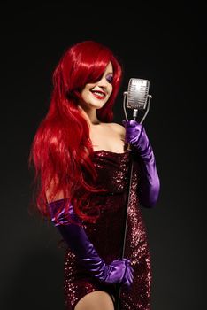 Young smile redhead woman with very long hair in red gown with microphone on the stand on a black background. Sexy gown on a beautiful slim figure. Long pink gloves