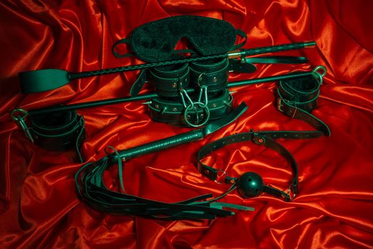 Bondage, kinky adult sex games, kink and BDSM lifestyle concept with a pair of leather handcuffs, flogger, ball gag and a coller with a leash attached on red silk with copy space