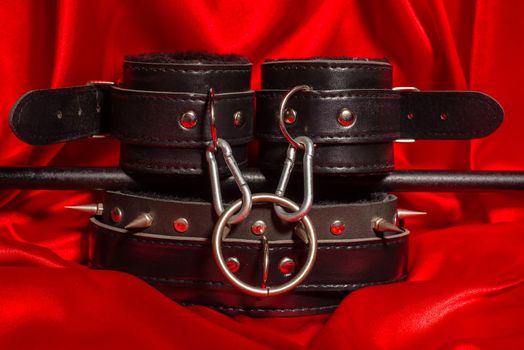 Close up bdsm outfit. Bondage, kinky adult sex games, kink and BDSM lifestyle concept with a pair of leather handcuffs, flogger, a leash attached on red silk with copy space.