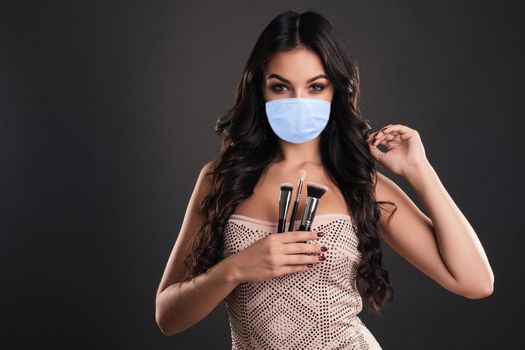 Portrait of the beautiful woman with make-up brushes near attractive face in mask. Adult girl posing over dark background. Coronavirus Outbreak Defense Concept