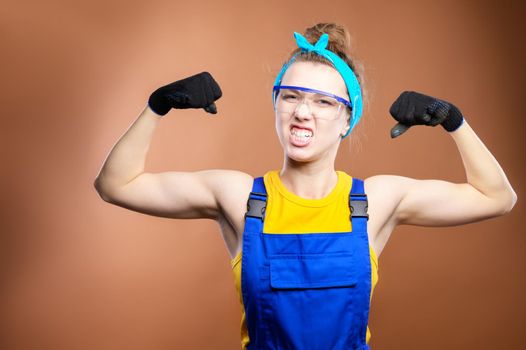 Young beautiful caucasian woman repairman worker with blue eyes in uniform showing arm muscles, proudly smiling and making a strong grimace. The concept of the rapid implementation of industrial projects. Literate contractors and wise construction superintendents