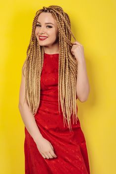 beautiful young woman dressed in a red dress, touches African pigtails. yellow background