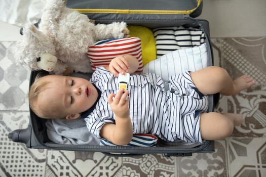 From above cute baby boy playing with toy car while lying in open suitcase near plush sheep before going on vacation