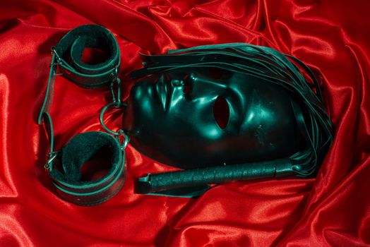 Bondage, kinky adult sex games, kink and BDSM lifestyle concept with a mask, pair of leather handcuffs, flogger, ball gag and a coller with a leash attached on red silk with copy space