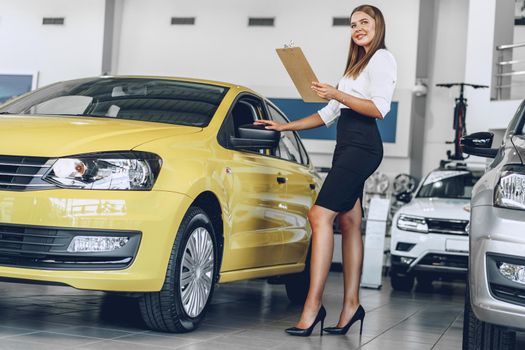 Attractive young female car dealer standing in showroom near a new car