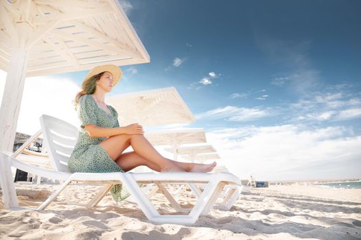 A blonde in a green dress sits on a sunbed and looks at the seascape under protective sunny wooden umbrellas. Young caucasian attractive woman by the sea on the beach.