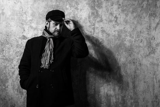 attractive young bearded man in a pea coat and cap is standing at the wall