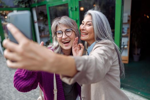 Positive senior Asian lady with grey haired friend take selfie standing on modern city street. Long-time friendship relationship