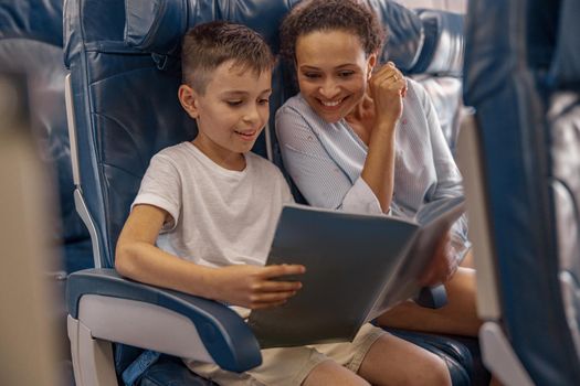 Boy, kid on board holding a book and looking trough it together with his mom during flight. Cabin crew providing service to family in airplane. Airline transportation and tourism concept