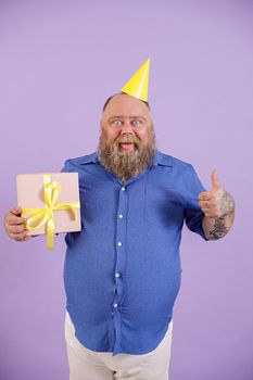 Funny bearded obese man with yellow party hat holds gift box showing thumb up and toungue on purple background in studio