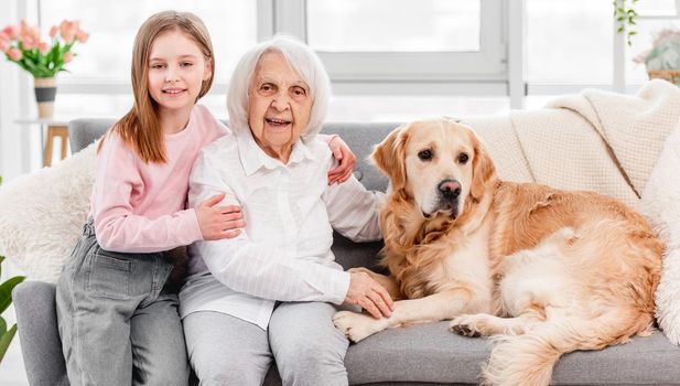 Family portrait of grandmother sitting on the sofa with granddaughter and golden retriever dog