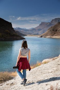 Back view a woman photographer in casual clothes with a professional camera stands on the shore of a lake in the mountains and looks at the rocks. Photo hikes and photo tours.