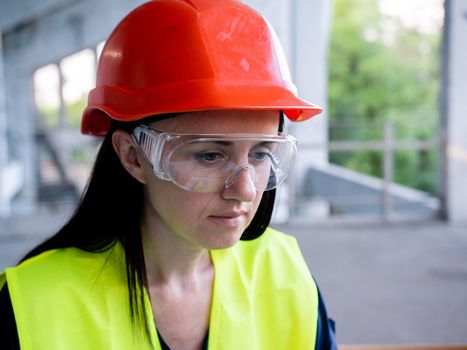 A female builder worker in an orange helmet sits on a construction site with a computer. The foreman checks the home renovation process.