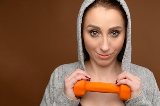 Portrait of a strong caucasian sporty woman in a tracksuit with a hood holds a fitness dumbbell in her hand and looks into the camera. Athletic lifestyle. Studio shot.
