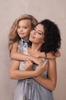 Little girl hugging happy mom from behind near grey wall