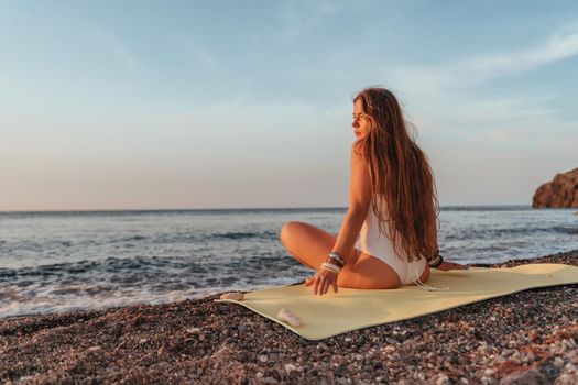 Young woman in swimsuit with long hair practicing stretching outdoors on yoga mat by the sea on a sunny day. Women's yoga fitness pilates routine. Healthy lifestyle, harmony and meditation concept.