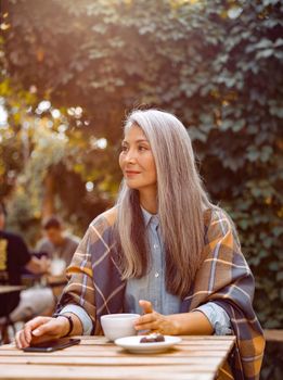 Pretty senior Asian woman with mobile phone and cup of coffee sits at small table on outdoors cafe terrace on autumn day