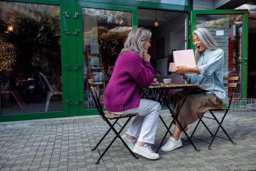 Excited senior Asian woman opens gift presented by best friend sitting at small table on outdoors cafe terrace on autumn day