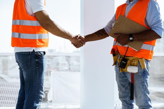 Two men engineers in workwear shaking hands against construction site, close up