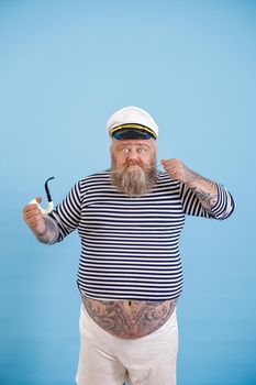 Funny bearded obese man wearing sailor suit with smoking pipe tightens up mustache on light blue background in studio