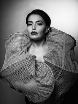 Young sexy slim tanned woman in a fishing cage posing against gray background. Black and white fashion portrait of beautiful girl with brunette hair.