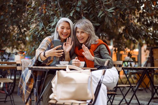 Positive mature women friends wave hands at videochat via smartphone sitting at small table in street cafe on nice autumn day