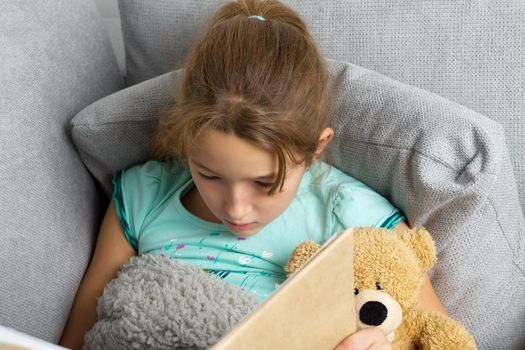 Beautiful girl sitting on comfortable sofa with teddy bear and reading book. Portrait of preteen girl with book and toy covered with plaid. Child resting on couch hugging favorite stuffed toy