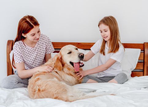 Two sisters sitting in the bed and petting golden retriever dog in the morning time. Girls with pet staying at home. Beautiful portrait of friendship between human and animal