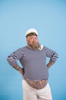 Confident bearded man with overweight in sailor suit with white cap and hands on waist stands on light blue background in studio
