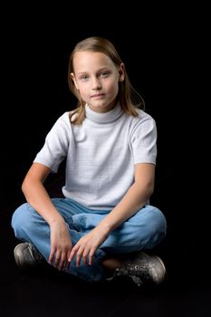 Girl sitting on floor with crossed legs. Stylish preteen blonde girl dressed gray jumper and jeans posing against black background. Portrait of preteen child looking seriously at camera