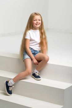 Girl in casual clothes sitting on white staircase. Cute smiling long haired girl wearing denim shorts, white t-shirt and sneakers posing in studio against white background