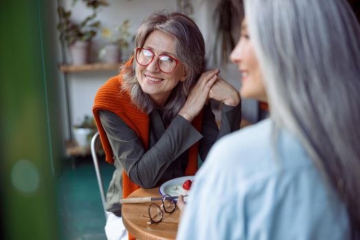 Positive mature woman with glasses and friend look through window sitting at small table in cozy cafe. Long-time friendship