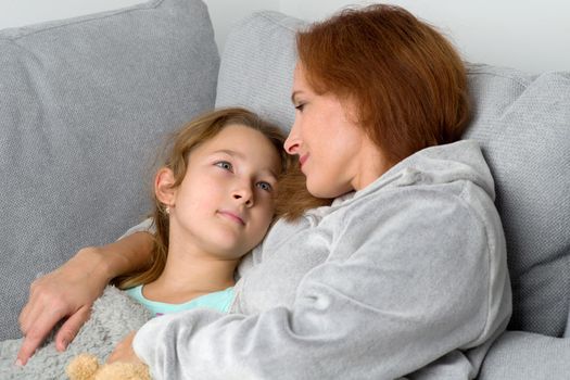 Mom and her daughter looking at each other. Loving mother hugging her preteen daughter. Cheerful woman and child wearing indoor clothes sitting together on couch at home