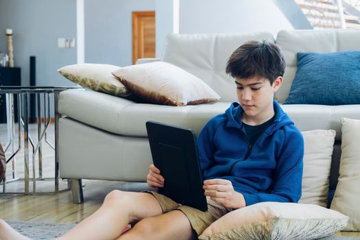 The boy using tablet computer while sitting on sofa at home. Lifestyle technology at home. Young person using telecommunications technology indoors, thoughtful.