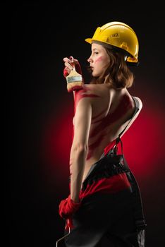 naked woman builder painter in overalls and construction helmet with a brush covered with thick red paint