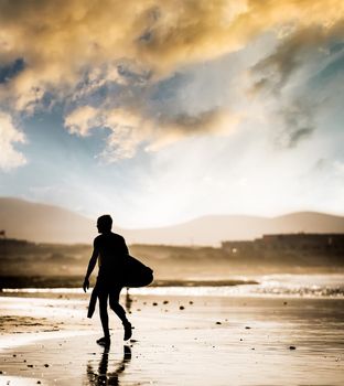 Silhouette of the man walking on the beach with the surfboard at sunset