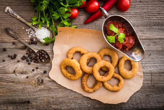 fried onion rings on parchment with sauce and vegetables on a wooden background