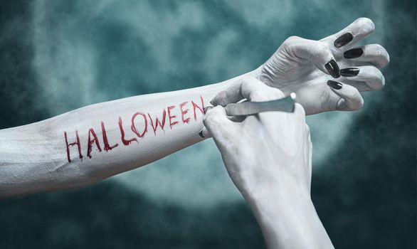 Unrecognizable dead woman writes the word Halloween by scalpel on her arm on background of full moon. View of human arms. Halloween or horror theme