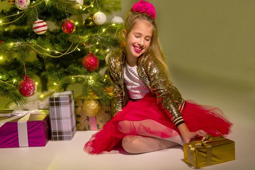Happy little girl rejoices at Christmas present. A girl sits near the shining lights of a Christmas tree which stands near a large panoramic window.