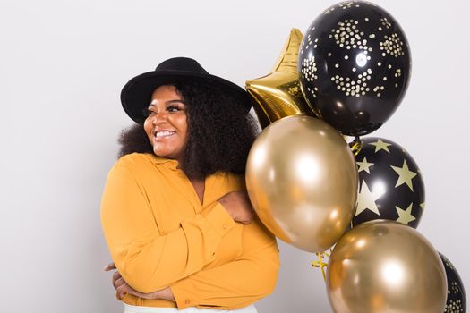 Portrait of smiling young African-American adult woman looking sweet on yellow background holding balloons.