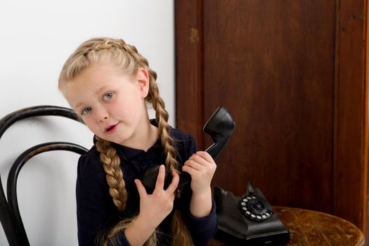 Blonde girl talking by old black phone. Lovely girl with braids sitting on chair in vintage room interior with old wooden furniture. Cute six years old kid speaking on retro telephone