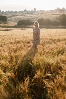 young girl in yellow outdoor dress stands at golden field of ears in rays of rising sun with her hair backlit and long shadow. grove and village in distance in background. Country side sunny morning