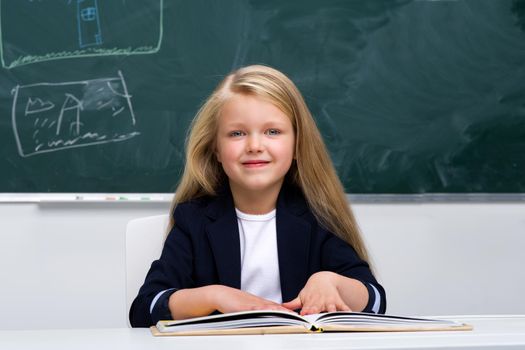Happy schoolgirl sitting at desk. Elementary school student in blue jacket and white blouse reading book on background of blackboard in classroom. Back to school, education concept