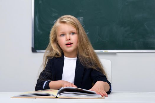 Happy schoolgirl sitting at desk. Elementary school student in blue jacket and white blouse reading book on background of blackboard in classroom. Back to school, education concept