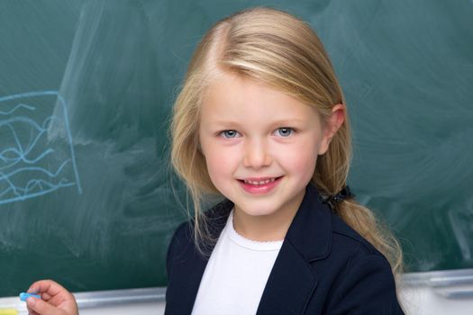 Portrait of cute happy schoolgirl. Beautiful blonde girl standing against green chalkboard in classroom. Cute elementary school student in blue jacket and white blouse