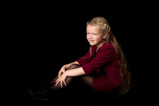 Happy long haired blonde girl. Adorable girl in stylish clothes sitting on floor hugging her knees. Lovely smiling little girl posing in studio against black background