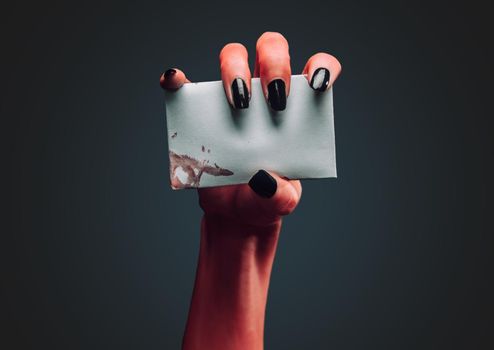 Red demon hand holds empty white card on dark background, space for text. Halloween or horror theme